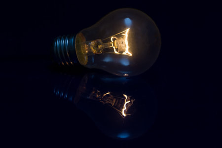 Light bulb with burning filament lay on blue shiny surface with reflectionの写真素材