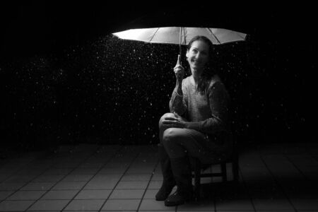 Beautiful woman with silver dress sitting in rain under an umbrella at nightの写真素材