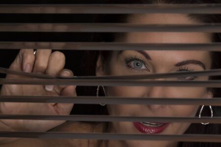 Portrait of a woman standing in the dark looking through blindsの写真素材