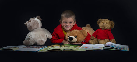 A smiling young boy with many stuffed animals reading a book before bed time on dark background
