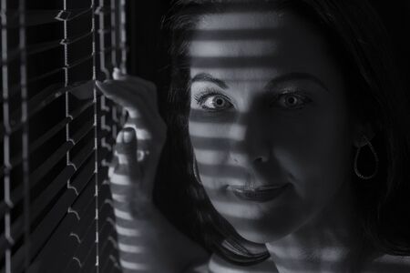 Portrait of a woman standing in the dark looking through blinds artistic conversionの写真素材