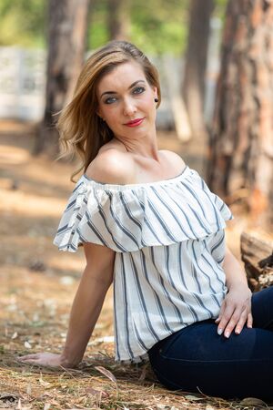 Portrait of a blonde woman with a striped blouse sitting on the ground in the forestの写真素材