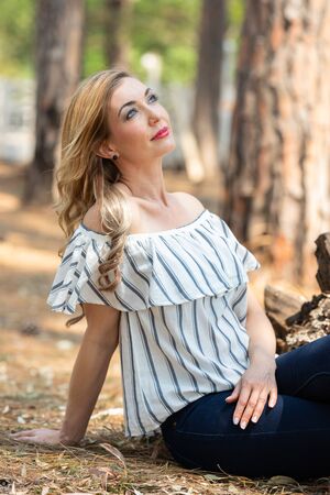 Portrait of a blonde woman with a striped blouse sitting on the ground in the forestの写真素材