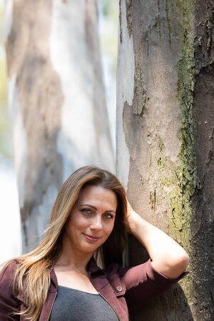 Portrait of a brunet woman with a blue denim and maroon jacket posing against a big treeの写真素材