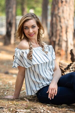 Portrait of a blonde woman with a striped blouse sitting on the ground in the forestの写真素材
