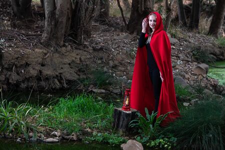 Attractive woman dressed a little red riding-hood cross a stream in a dark forest with a lanternの写真素材