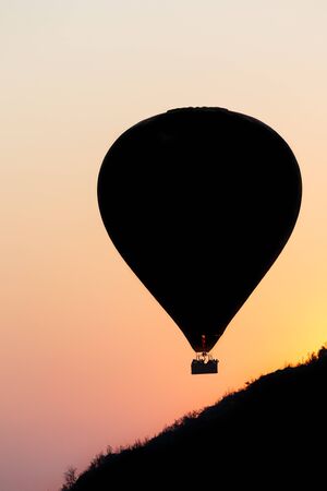 One hot air balloon floating over low hills into an orange sunsetの写真素材