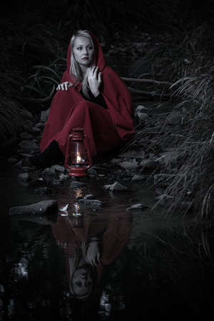 Attractive woman dressed a little red riding-hood cross a stream in a dark forest with a lanternの写真素材