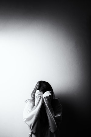 Black and white image of a woman covering her face with long shirt sleeves, leaning against a wall, symbolizing sadness, anxiety, and emotional painの写真素材