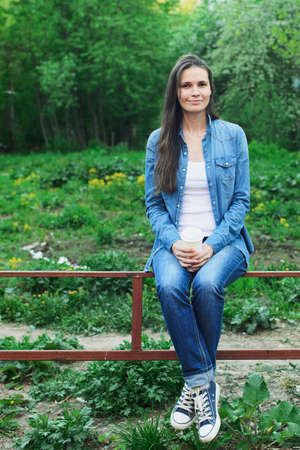 Young woman with cup of coffee sitting on a metal fence. jean lifestyleの写真素材