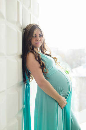Pregnant girl standing against wall in azure dress near windowの写真素材