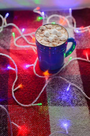 blue ceramic cup of hot chocolate with marshmallow and multi colored garland on a red checkered blanketの写真素材