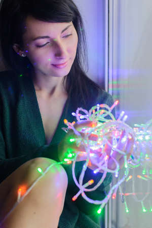 young woman entwined colorful festive garland sitting on window sillの写真素材