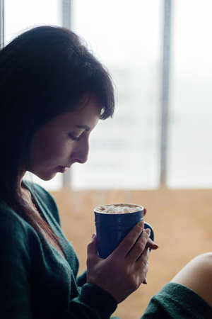 brunette young woman in green sweater sitting on window sill with cup of cocoaの写真素材
