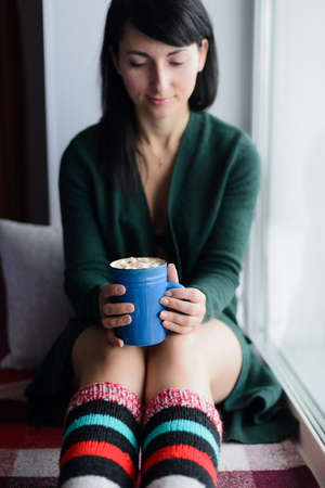 brunette young woman in green sweater sitting on window sill with cup of cocoaの写真素材