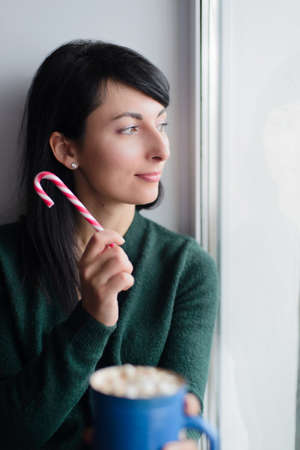 brunette young woman in green sweater sitting on window sill with cup of cocoaの写真素材
