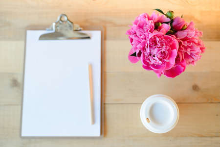 Top view flat lay of blank clipboard with pencil, cup of coffee and pink peony bouquet on wooden backgroundの写真素材