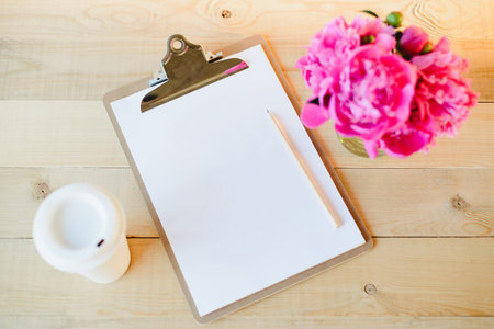 Top view flat lay of blank clipboard with pencil, cup of coffee and pink peony bouquet on wooden...