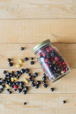 Red, white, black currant, red and black raspberries, white strawberries in glass jar and sprinkle on wooden table as  canned foodの写真素材
