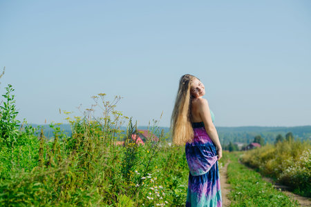 white caucasian blonde young woman with colorful long dress going on road in fieldの写真素材