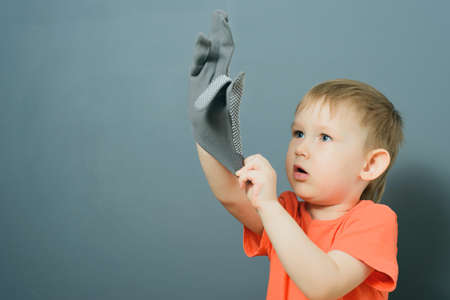 Caucasian blond baby boy in orange t-shirt puts on tissue gloves on his hands on gray background wallの写真素材