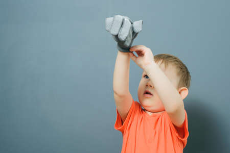 Caucasian blond baby boy in orange t-shirt puts on tissue gloves on his hands on gray background wallの写真素材