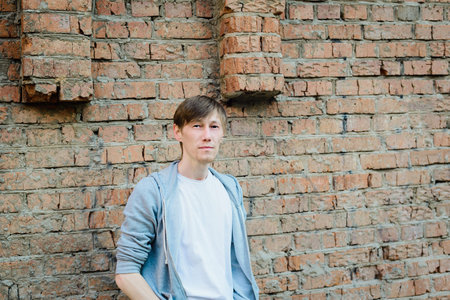 young 30s age man in white t-shirt and gray hoodie stands on old grange red brick wall background,...