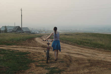 white caucasian young woman in casual clothing walking with bike, looking on village in evening fog, view from back in full body size, lifestyles stock photo imageの写真素材