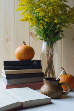 autumn still life with books stacked, orange pumpkins, clay drinking cup with spoon, solidago bouquet, side view of vertical stock photo imageの写真素材