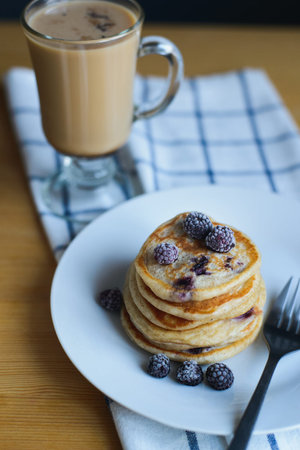 small circular fritter or pancakes with blackberry, frozen berries, fork on plate and coffee on...