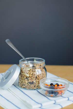 baked muesli with nuts and oat flakes, sea buckthorn and blueberry berries in glass bowl on striped textile napkin on wooden table, close up view from above of vertical still life stock photo imageの写真素材