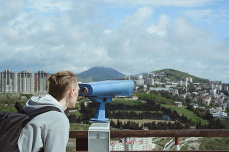 young caucasian hipster man looking through stationary binoculars at sea, mountains and city on observation deck, horizontal outdoors stock photo imageの写真素材