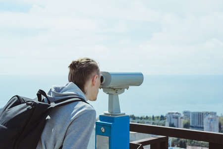 young hipster tourist man looking through metal coin operated binoculars on sea, sky and city, horizontal lifestyle stock photo imageの写真素材