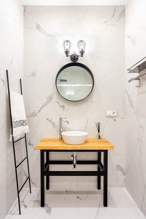 Modern style bathroom with gray and white tiles. There is a large Round mirror with luminous lamps,...