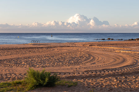 Calm morning at empty beach with a car tyre prints on sand, with sea and clouds at the background