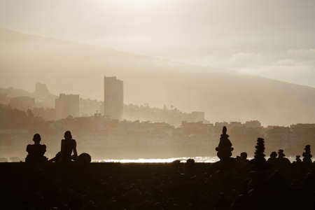 People are sitting on the beach on a sunny evening and looking at the sea. In the background the coast and buildings in a haze of water. Tenerife. Puerto de la Cruz. Playa Jardin.の写真素材