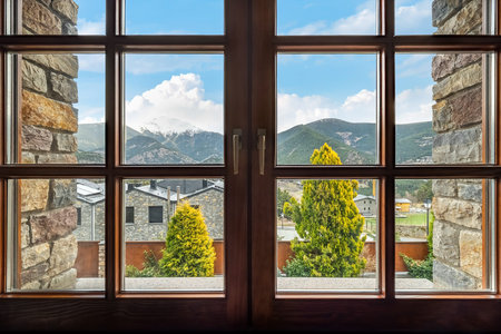 Window with a view to the ridge of mountains, snow-capped peak, clouds, sky, trees and houses in Andorraの写真素材