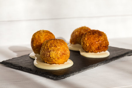 Fried meat balls or croquettes with white sauce on black stone plate on white wooden background....