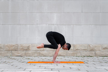 Latin man practicing yoga outdoor in a city, standing in crane pose on orange mat, with gray wall at...
