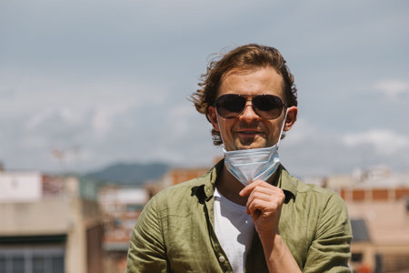 Young beautiful man takes off protective medical mask from face. Happy and smiling positive guy in...
