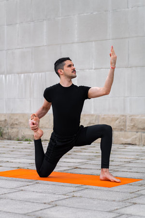 Handsome sporty latin man exercising in a city, doing yoga, sitting in variation of One Legged King...