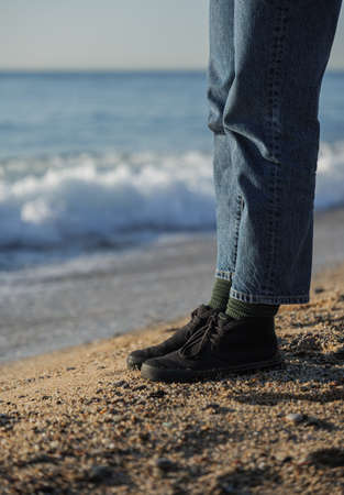 Legs in blue jeans and black shoes standing on the beach with sea waves at the backgroundの写真素材