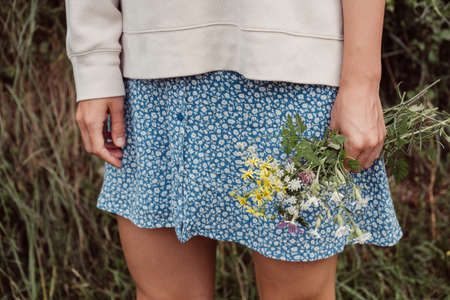 A countryside girl in a blue skirt and a white blouse stands with wildflowers in her hand.の写真素材