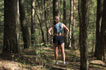 Rear view of female trail runner dressed in sportswear standing on trail in the forest and preparing...