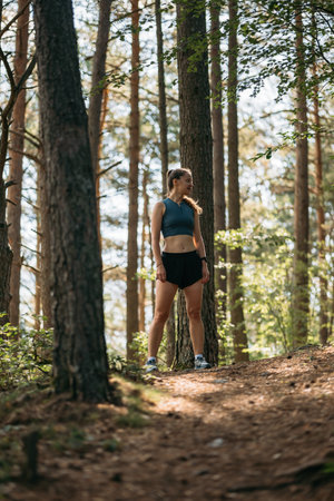 Young woman runner dressed in sportswear standing on trail in green forest, ready to run while preparing for marathonの写真素材