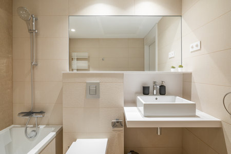 Cozy bathroom in modern design with huge mirror on the wall and bright lighting. Walls are in beige tiles and white sanitary ware. Room for taking morning shower and relaxing evening baths.の写真素材