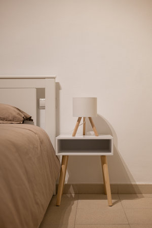 Minimalist bedroom with taupe bedding, white headboard, wooden nightstand and small tripod lampの写真素材
