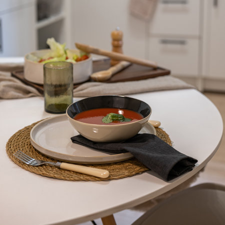 Table setting with bowl of tomato soup on ceramic plate black napkin fork and salad dish in backgroundの写真素材