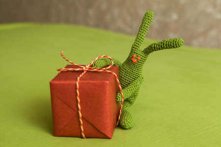 Green crochet bunny hugging red gift box tied with rope bow placed on green surface in cozy indoor settingの写真素材