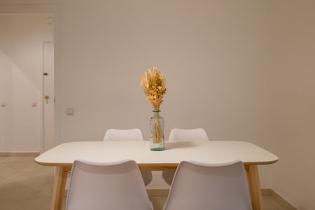 Minimalist dining area with white table, four white chairs and dried flowers centerpiece in a glass...